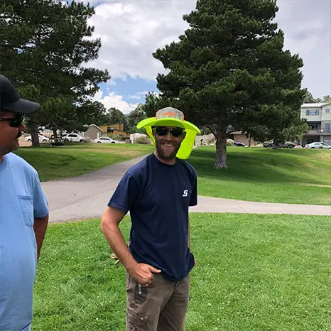 Los Alamos Landscaping team member smiling at an outdoor gathering, wearing sunglasses and a neon safety vest draped over his hat.