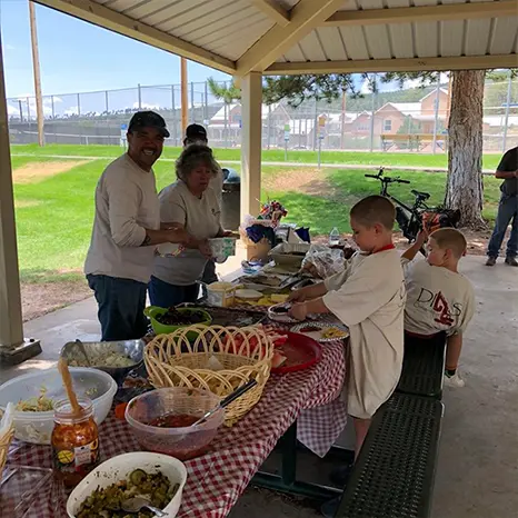 Los Alamos Landscaping team and community members enjoying a picnic under a pavilion, with a table full of food and children serving themselves.