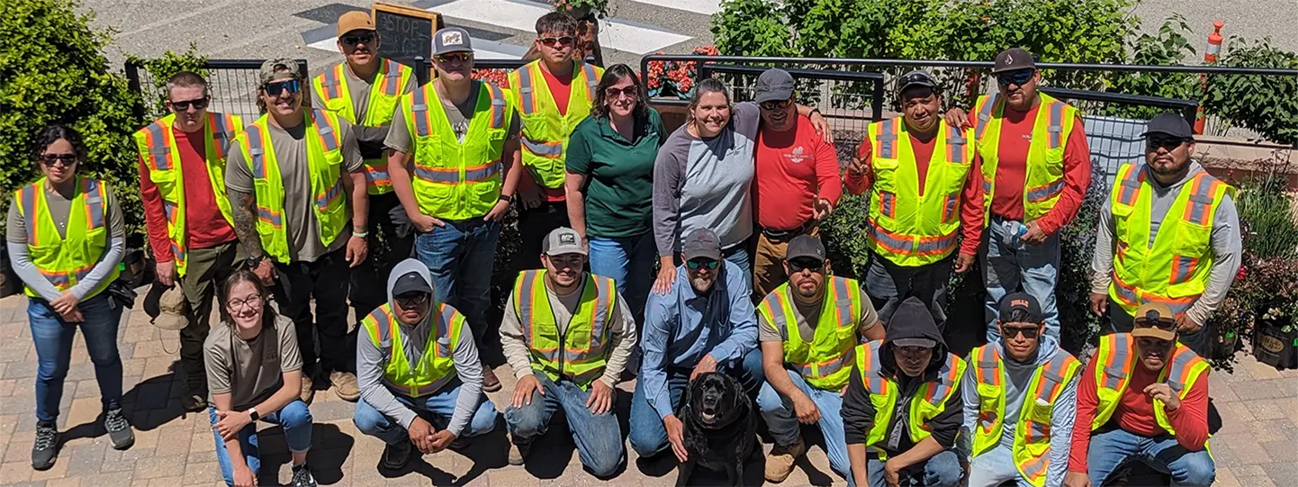 Los Alamos Landscaping team posing together outdoors, wearing safety vests and work gear, with a black lab at the center.