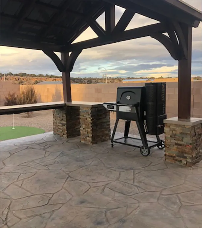 A custom outdoor pavilion featuring a wooden pergola, stacked stone bar, and a barbecue station on a stamped concrete patio, set against a scenic desert landscape.